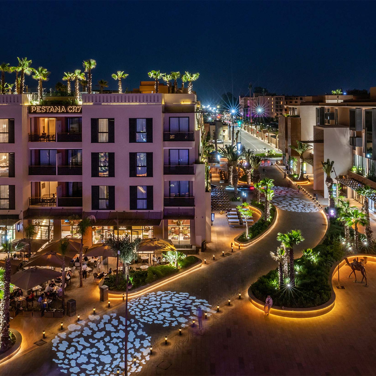 Exterior de noche del Pestana CR7 Marrakech, ubicado en la M Avenue, con varias tiendas, palmeras y arbustos iluminados