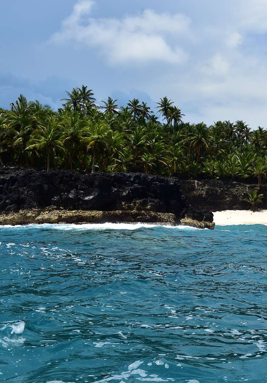 Vista desde un pequeño barco azul navegando hacia la costa rocosa con palmeras en Ilhéu das Rolas
