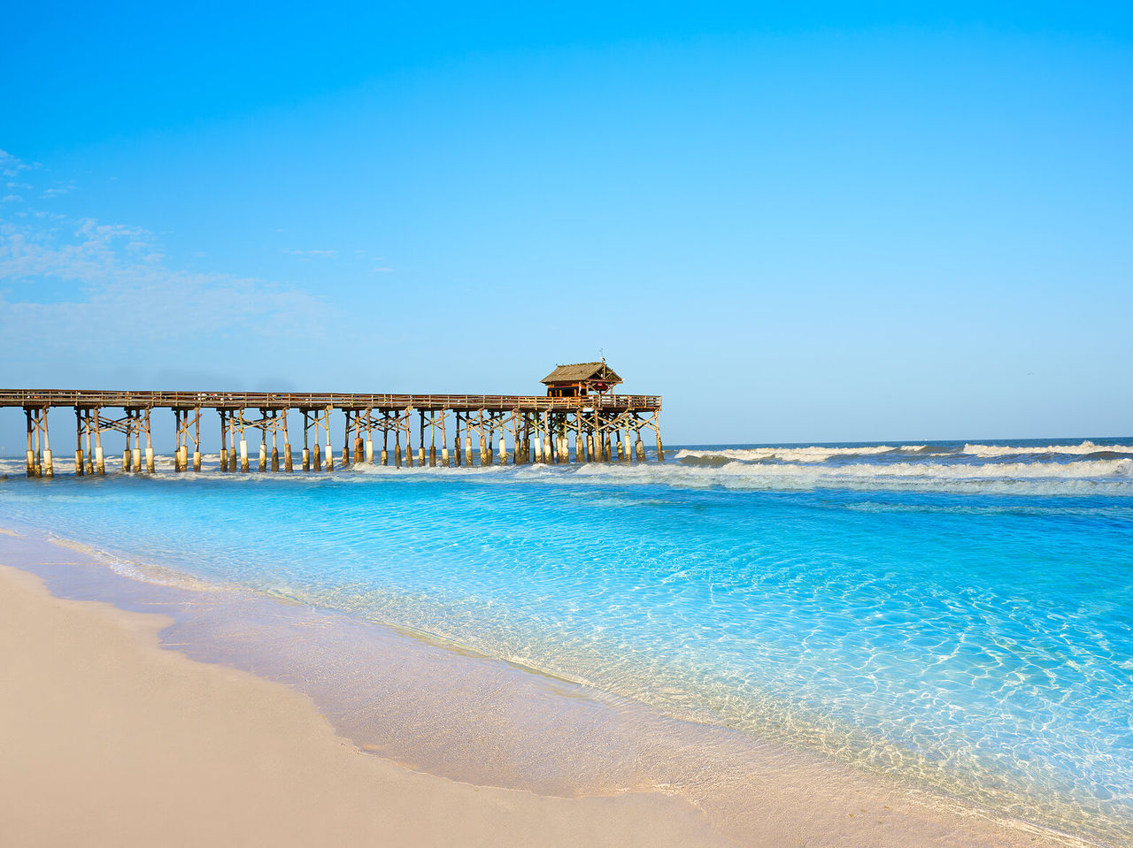 Largo muelle de madera sobre agua cristalina, ofreciendo una vista de una playa de arena blanca con un mar relajante