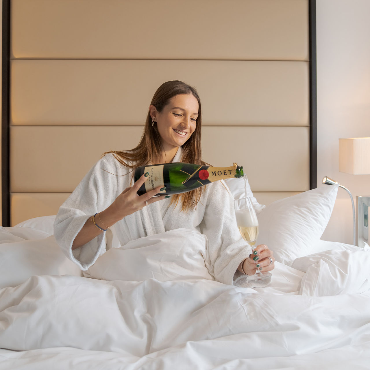 Chica sonriendo sentada en la cama con un albornoz blanco y una botella de champán sirviendo en la copa