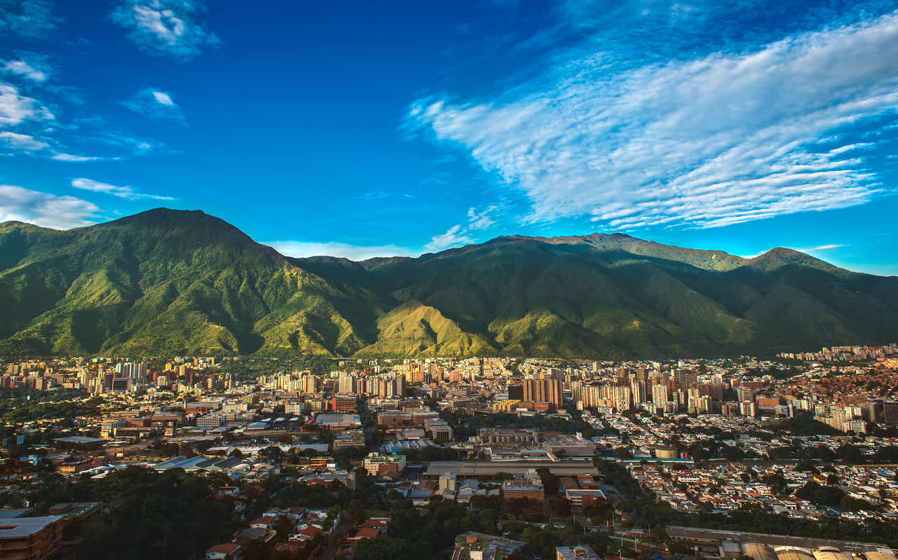 Vista aérea da cidade de Caracas, com prédios altos, contrastando com a natureza montanhosa, e o céu azul