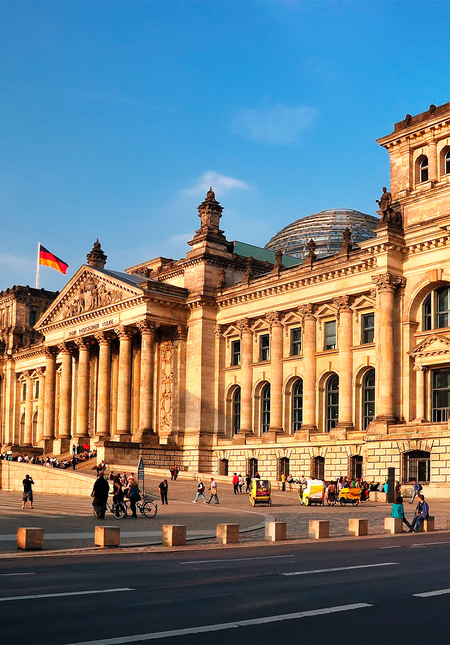 Edifício do Reichstag em Berlim ao entardecer, com bandeiras da Alemanha hasteadas e pessoas caminhando na frente