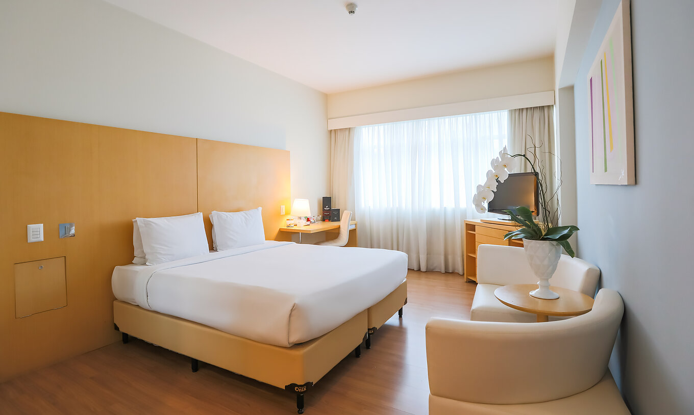 The Queen Standard at Pestana Curitiba has a room with a double bed, sofas, a desk, and a furniture unit with a TV