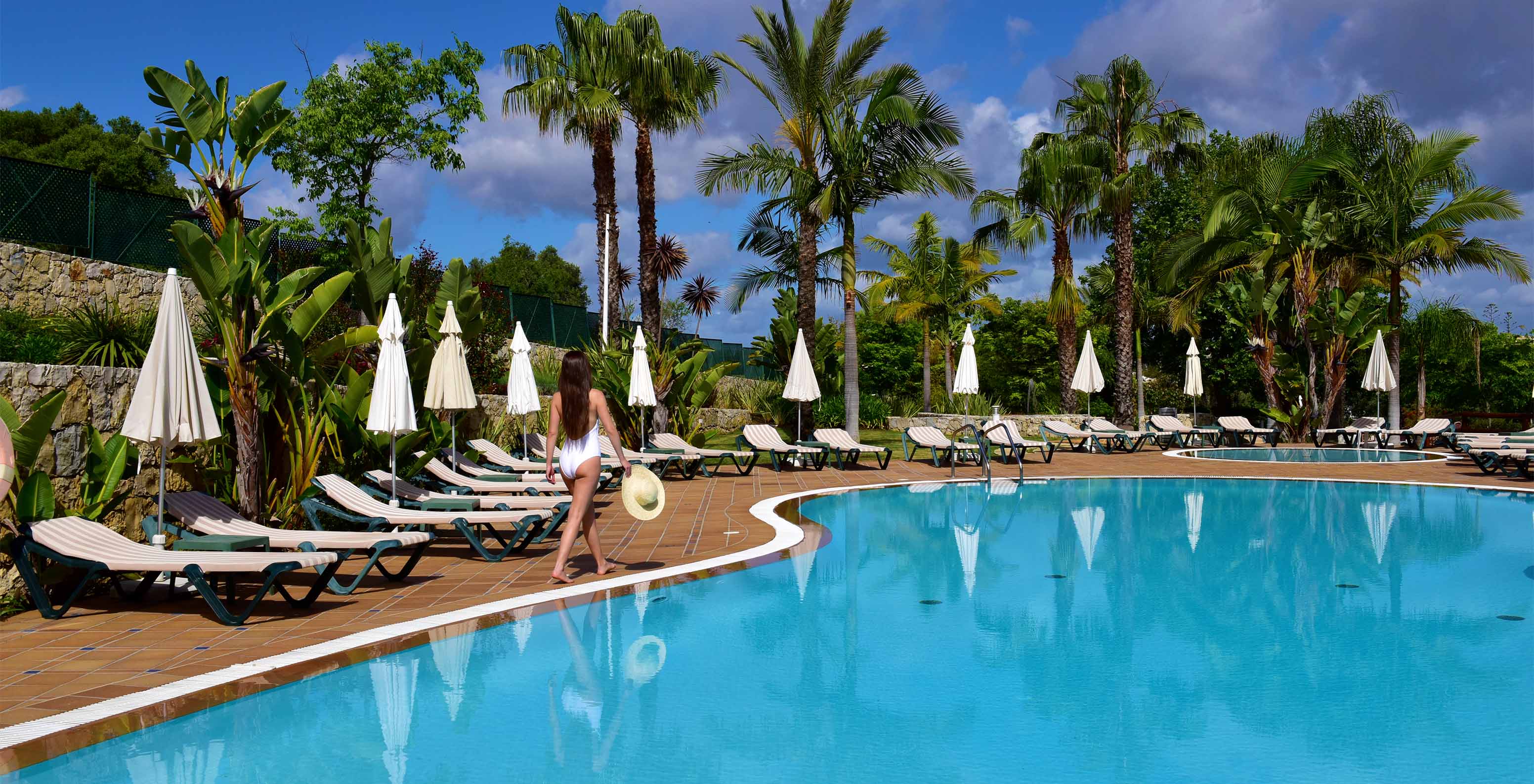 Outdoor pools with sun loungers, umbrellas, and palms, with a lady walking in a white swimsuit