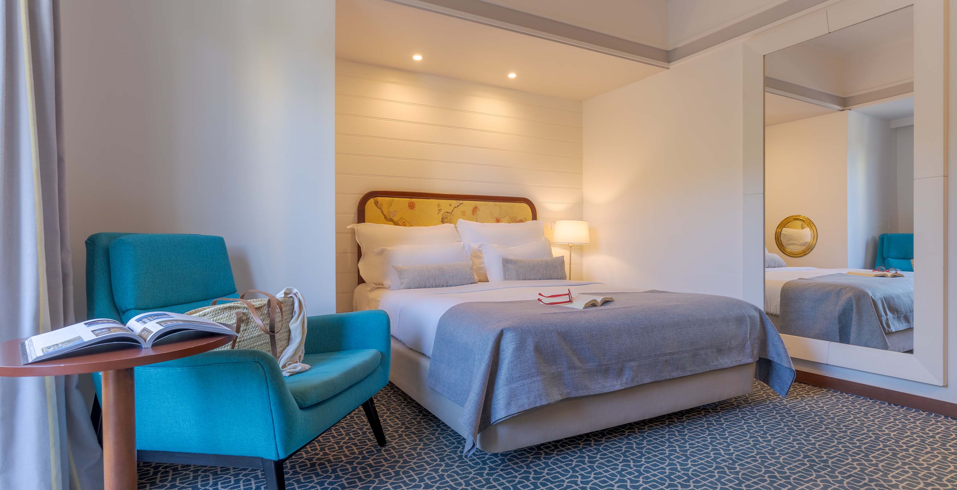The Deluxe at Pestana Alvor Praia has a double bed, a blue armchair, a mirror, and a geometric rug