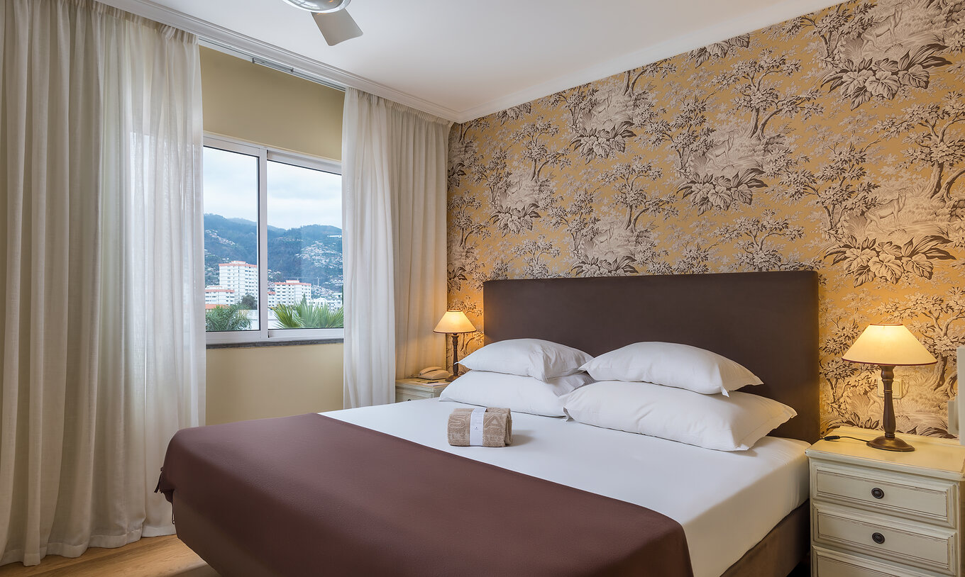 The Superior Family room at Pestana Village has a double bed with headboard and brown bedspread, floral wallpaper and balcony