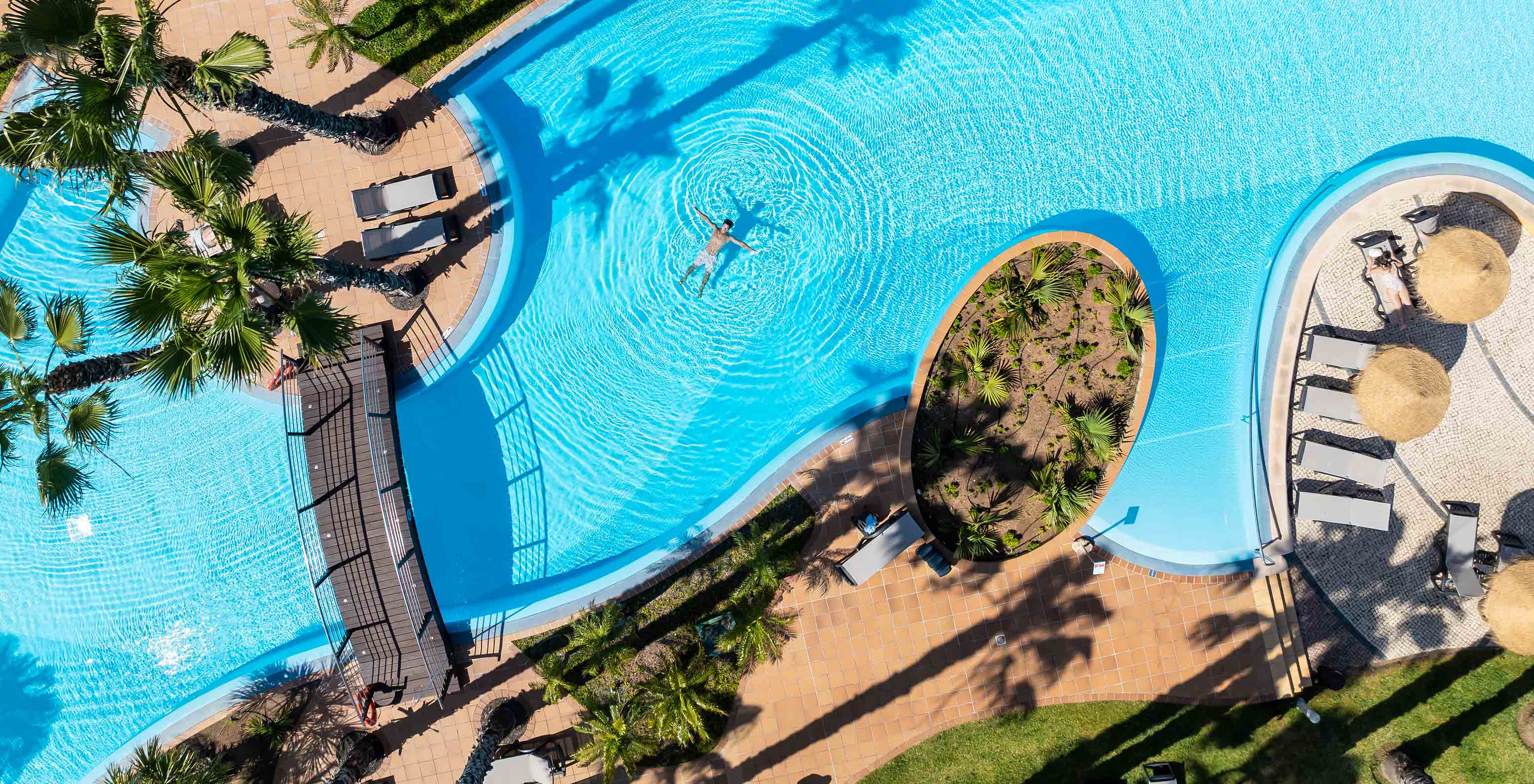 View of the outdoor pool at Pestana Porto Santo, with a guest in the water, a bridge, and tropical palm trees