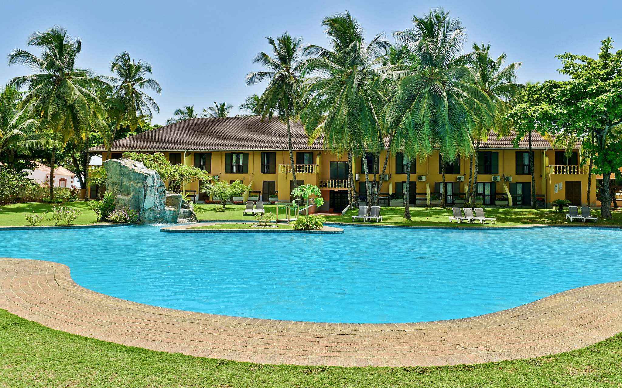 View of the yellow building with several windows, outdoor pool, palm trees, and sun loungers of a 4-star hotel in São Tomé
