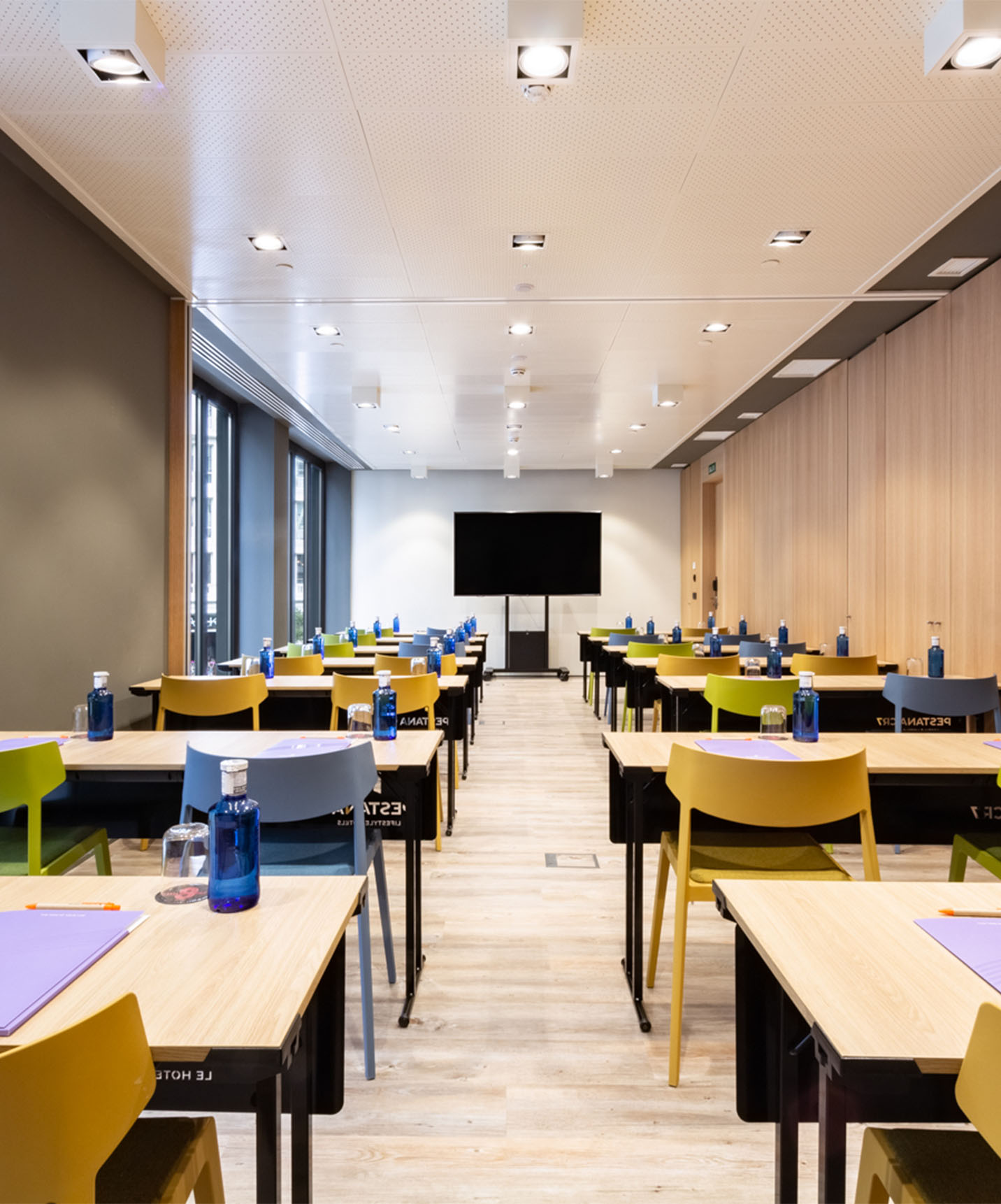Event and meeting room with wooden tables and yellow, green, and blue chairs, with TV and wooden walls