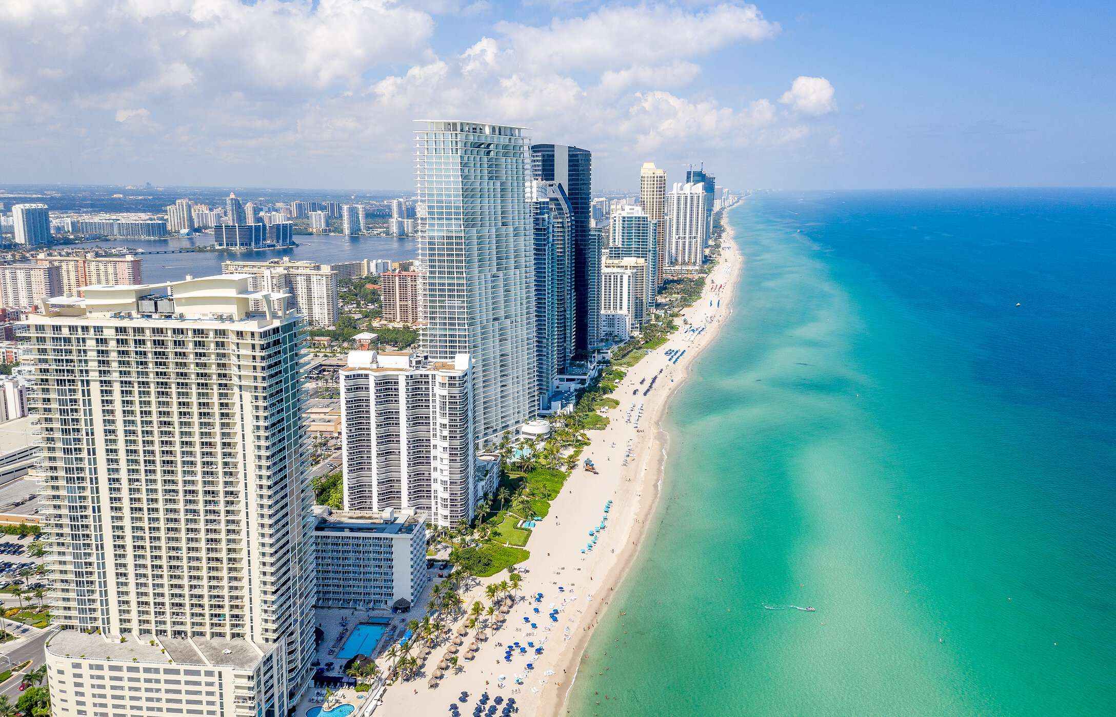 Aerial view of the coast of Miami Beach, Florida, with a long stretch of white sand, crystal-clear water, and tall buildings