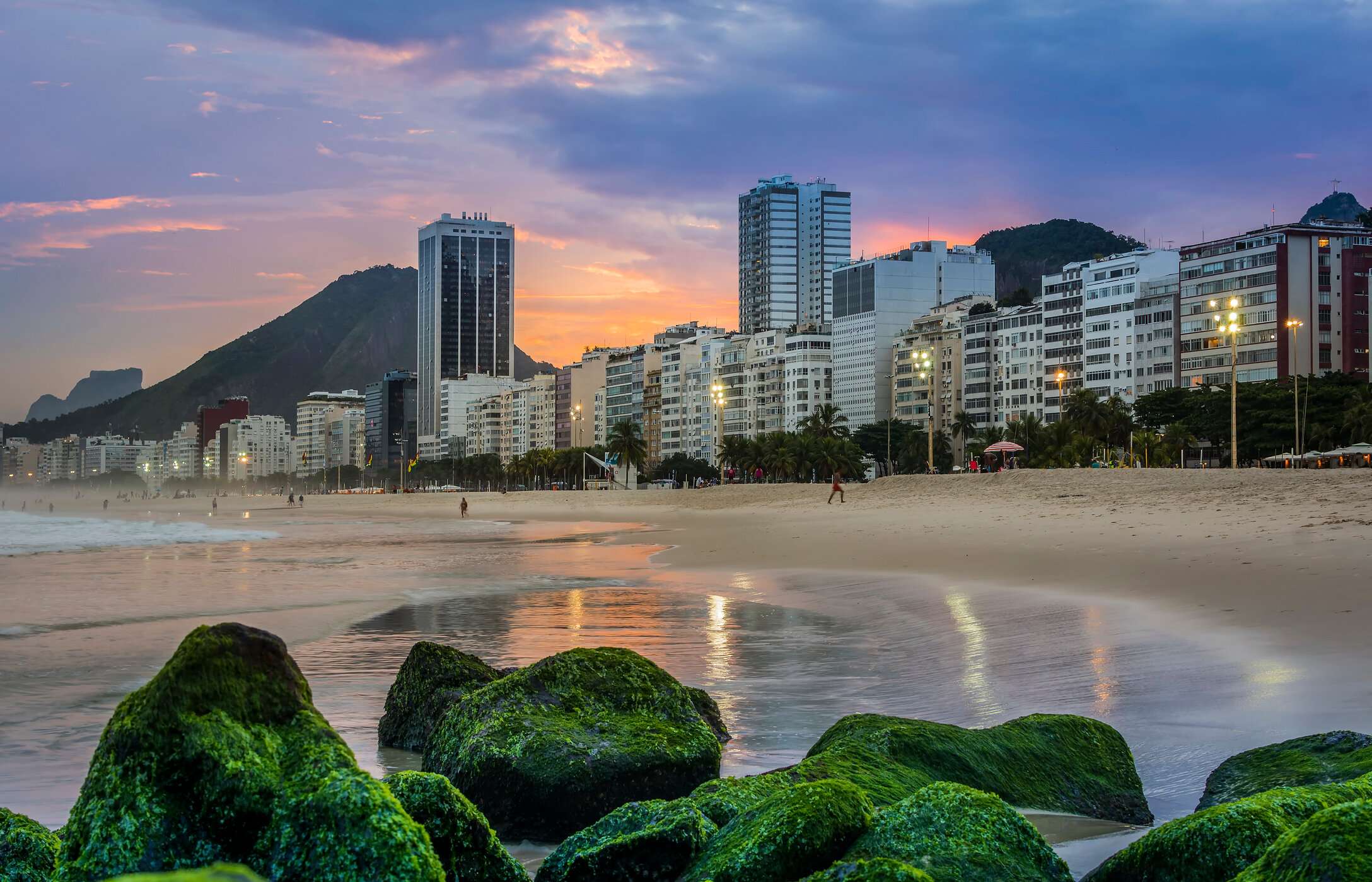 Playa de Copacabana, con arenas doradas, olas del mar y un paseo marítimo concurrido, rodeado de edificios y montañas al fondo.
