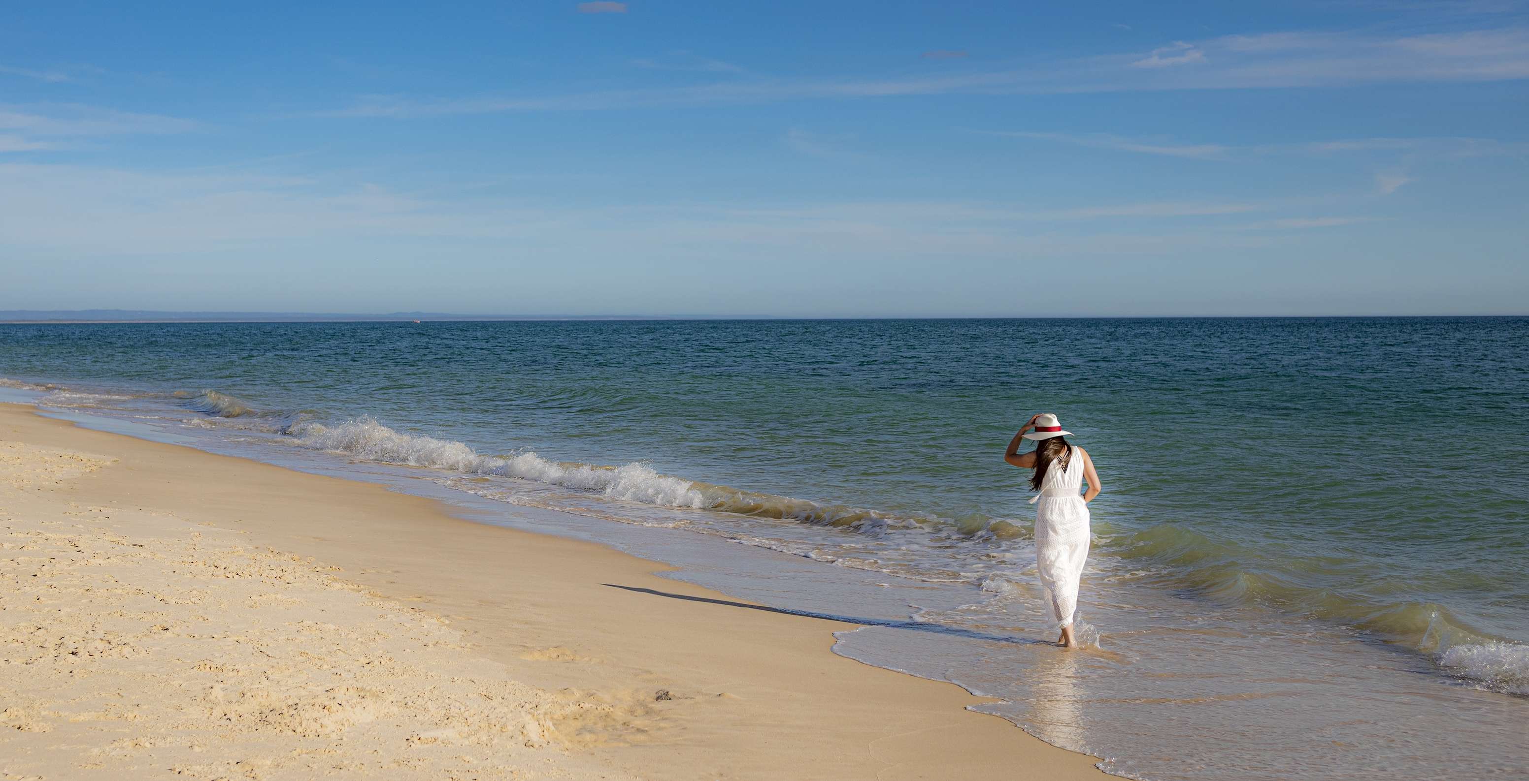 Mujer con vestido y sombrero blanco pasea junto al mar cerca del Pestana Tróia Eco-Resort, con acceso directo a la playa