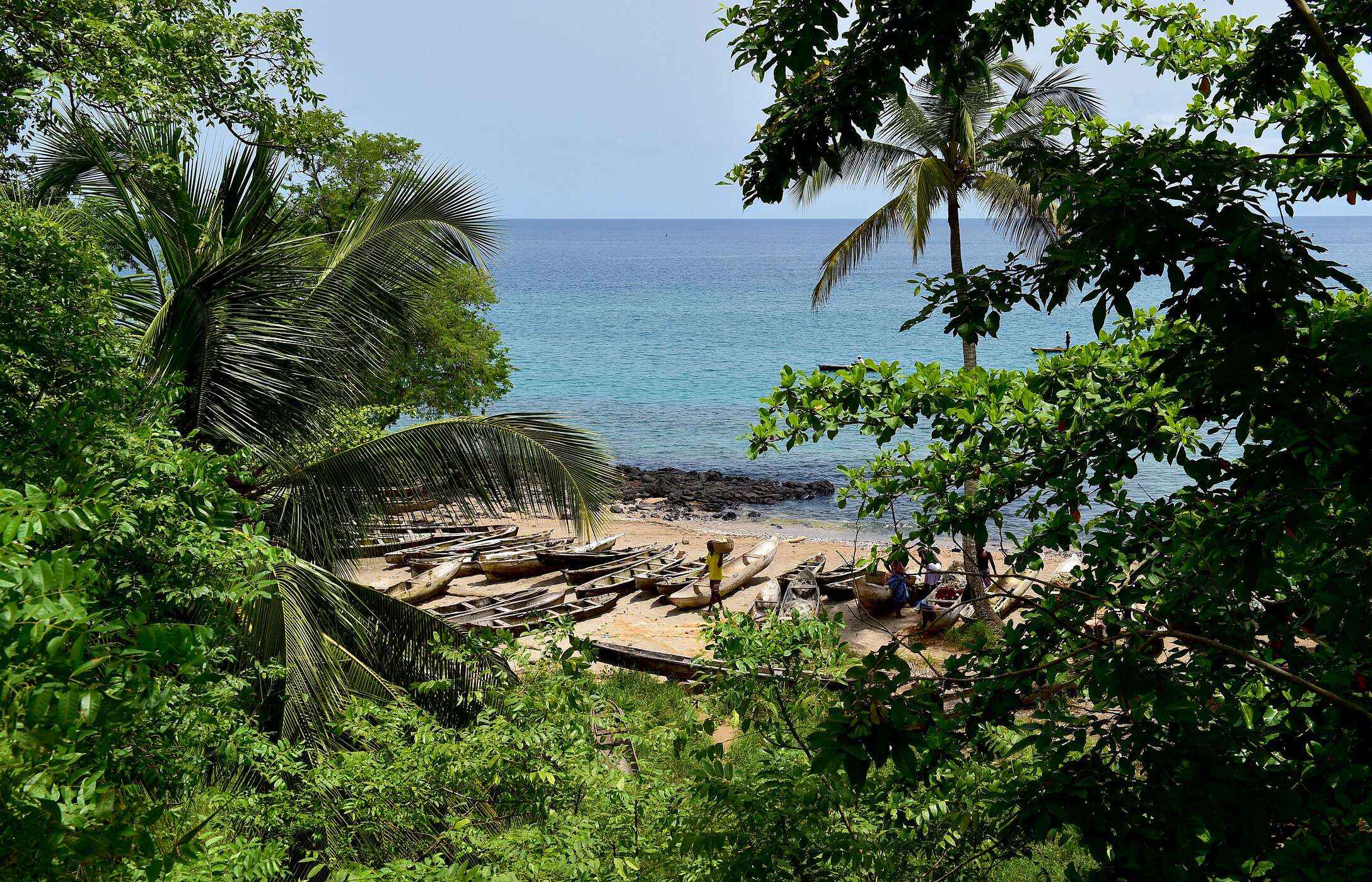 Uitzicht op een strand in São Tomé en Príncipe, omgeven door vegetatie, met kano's op het zand en de zee op de achtergrond.