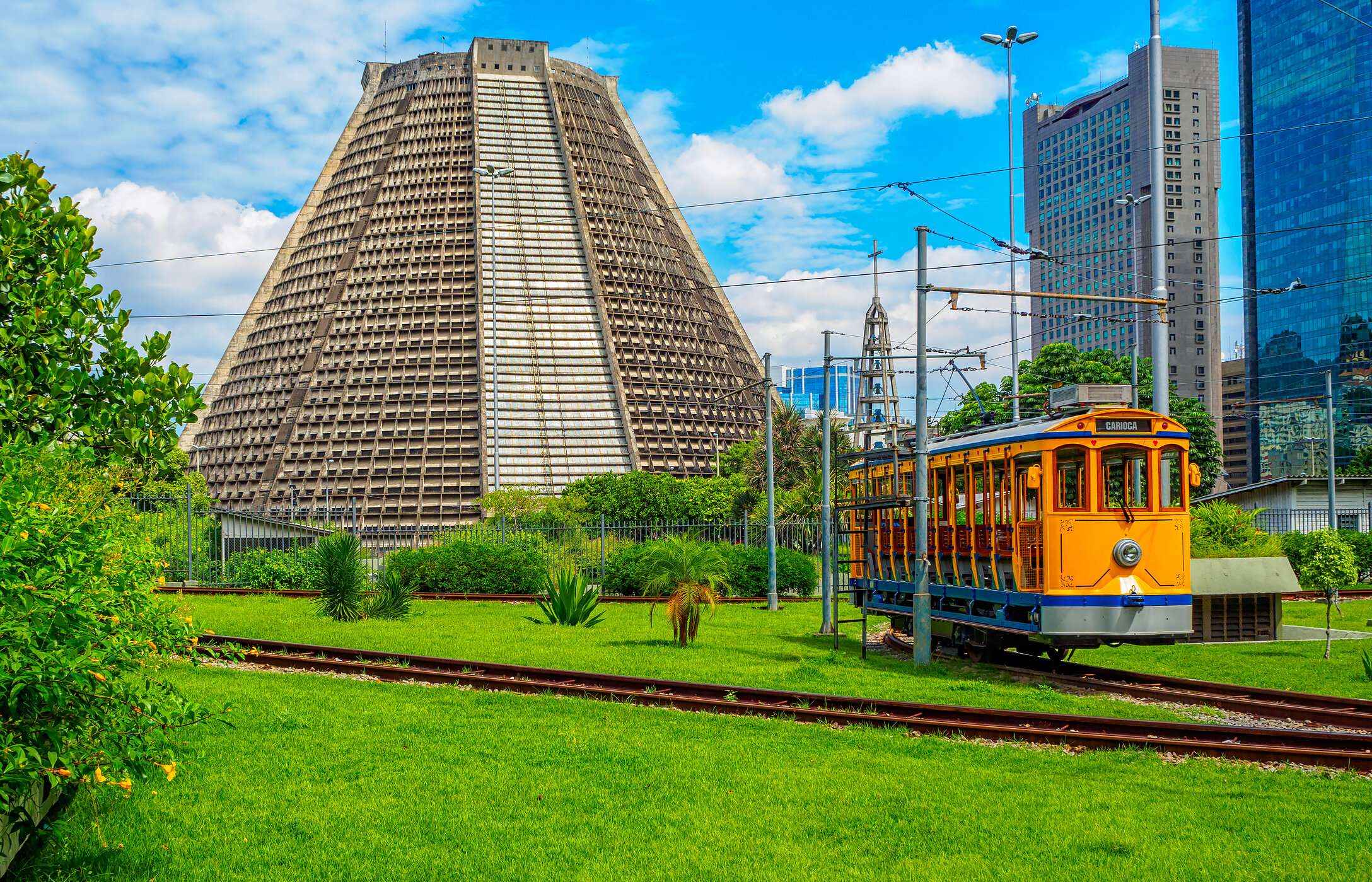 Catedral Metropolitana do Rio de janeiro, rodeada por paisagem verde, um elétrico antigo e, prédios em segundo plano