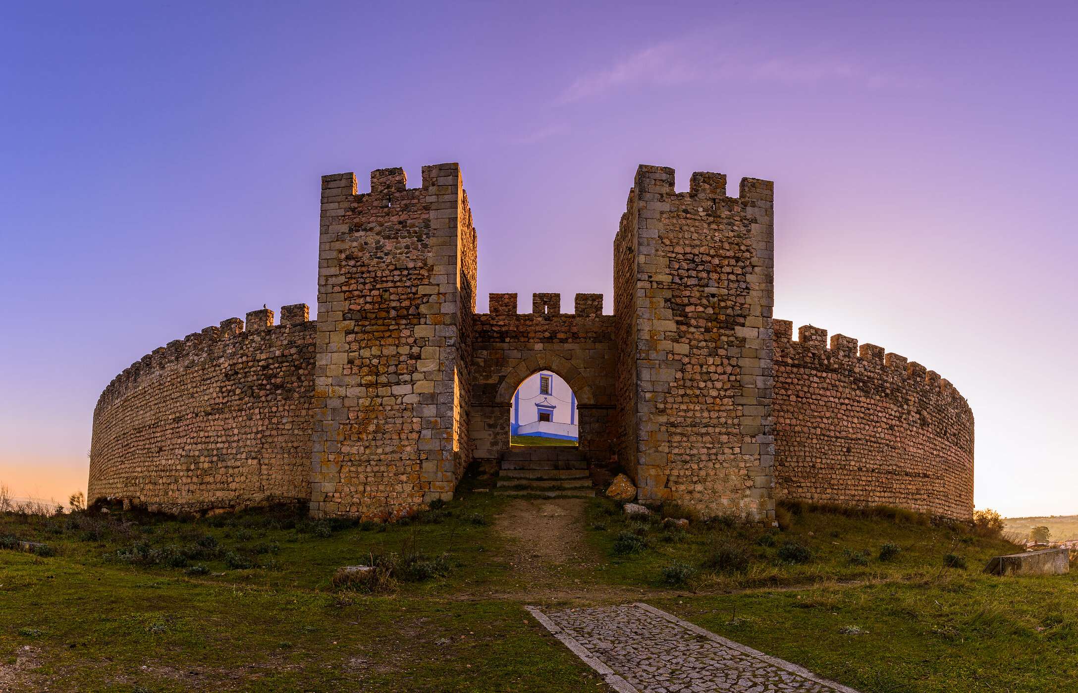 Vista frontal imponente da entrada do castelo de Arraiolos, com as muralhas de pedra e vegetação à volta