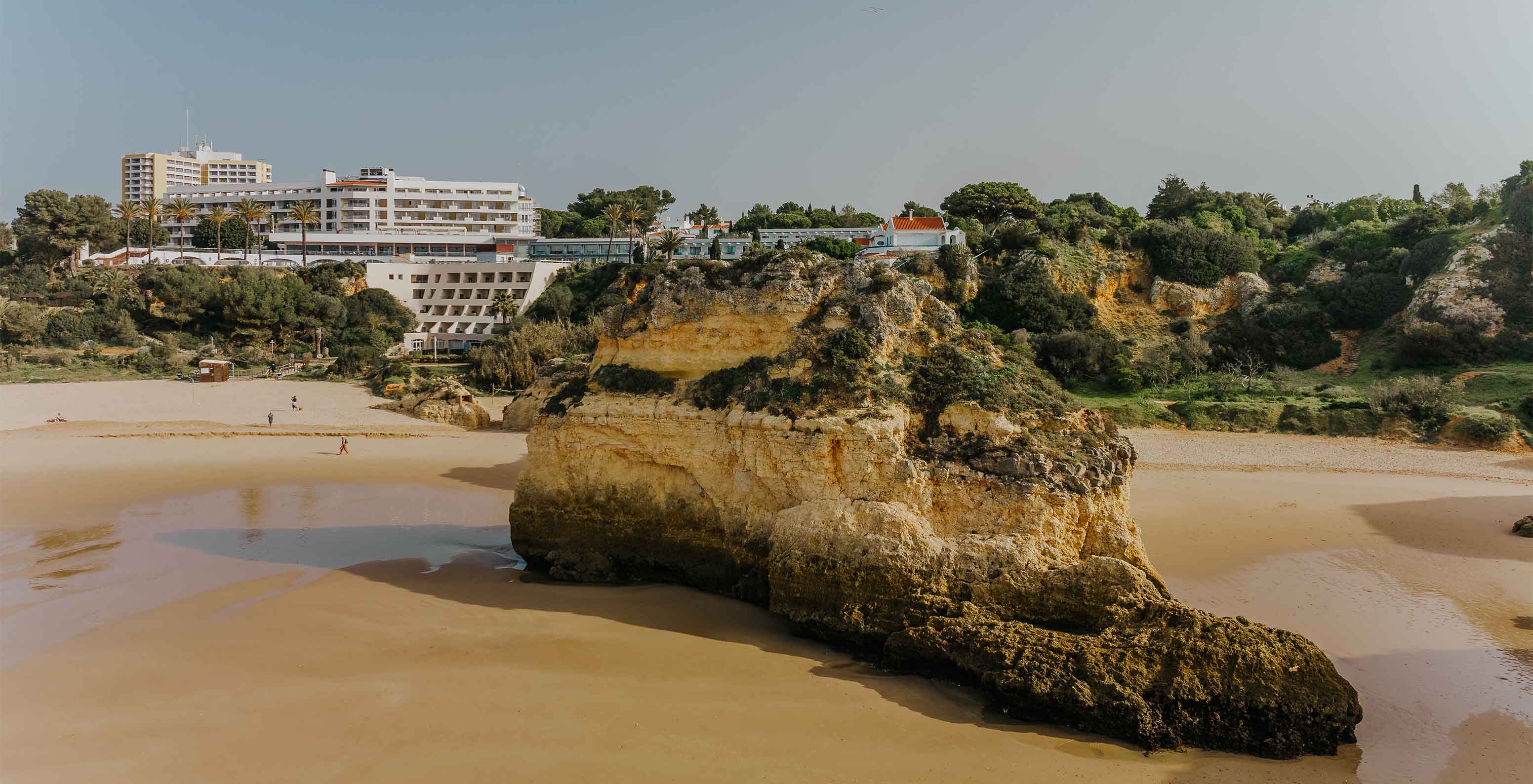 Praia do Alvor com rochas, areia, vegetação, a falésia e edifícios no fundo, com céu limpo