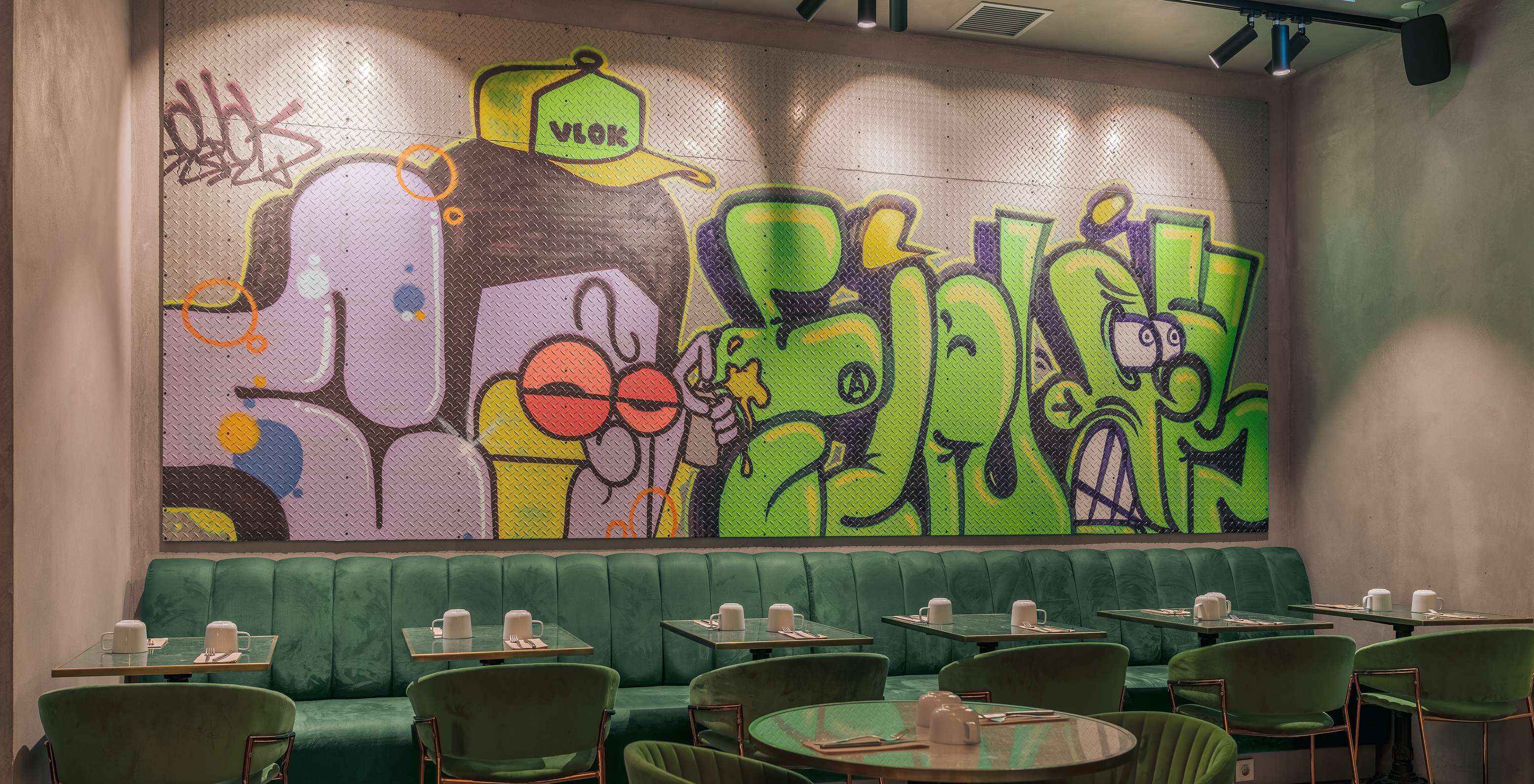 Restaurante com mesas, cadeiras e sofás verdes, com copos de loiça branca e com uma parede colorida com grafites
