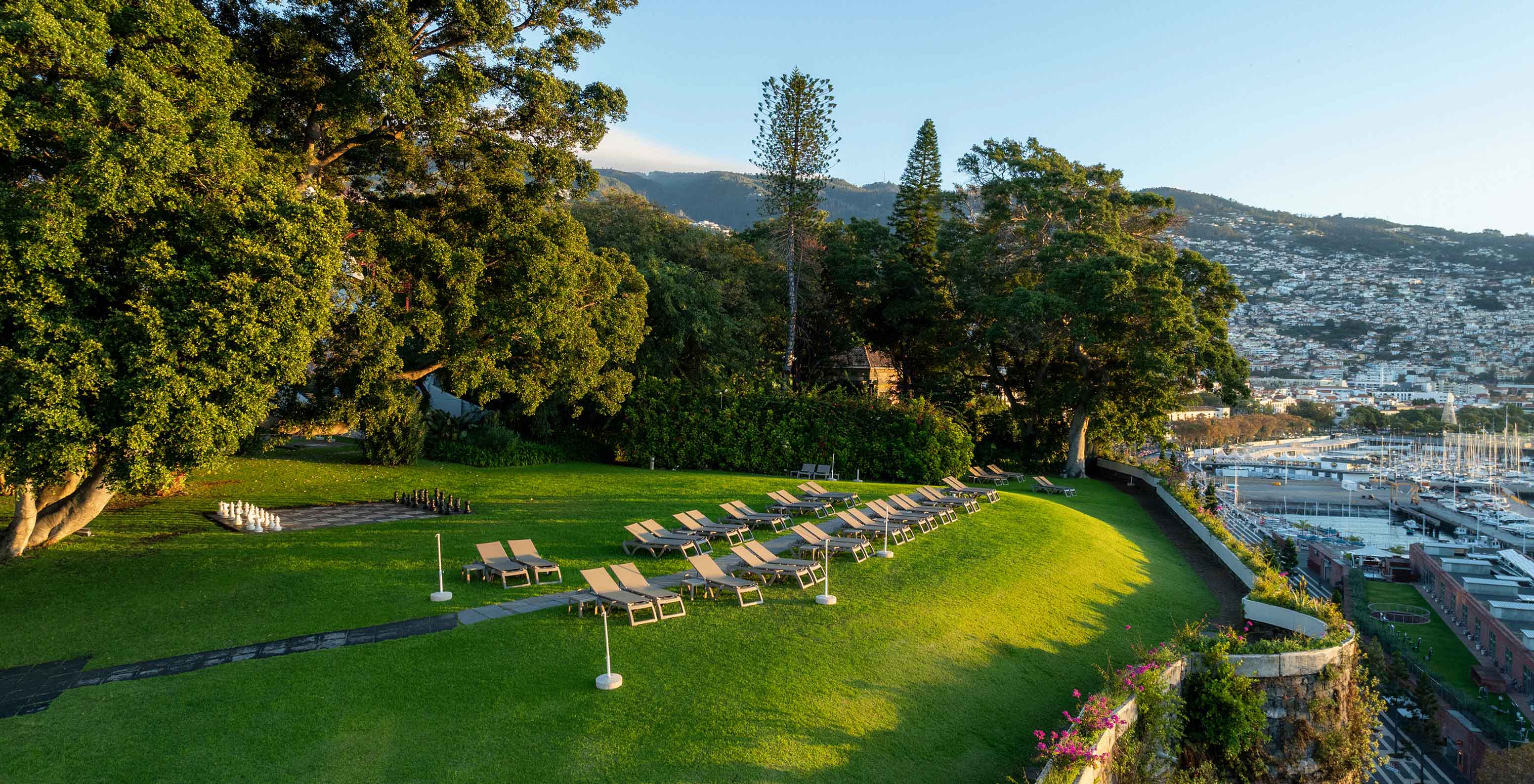 Vista do jardim do Pestana Casino Park, durante o pôr do sol, com uma cidade ao fundo e um cruzeiro ancorado no mar