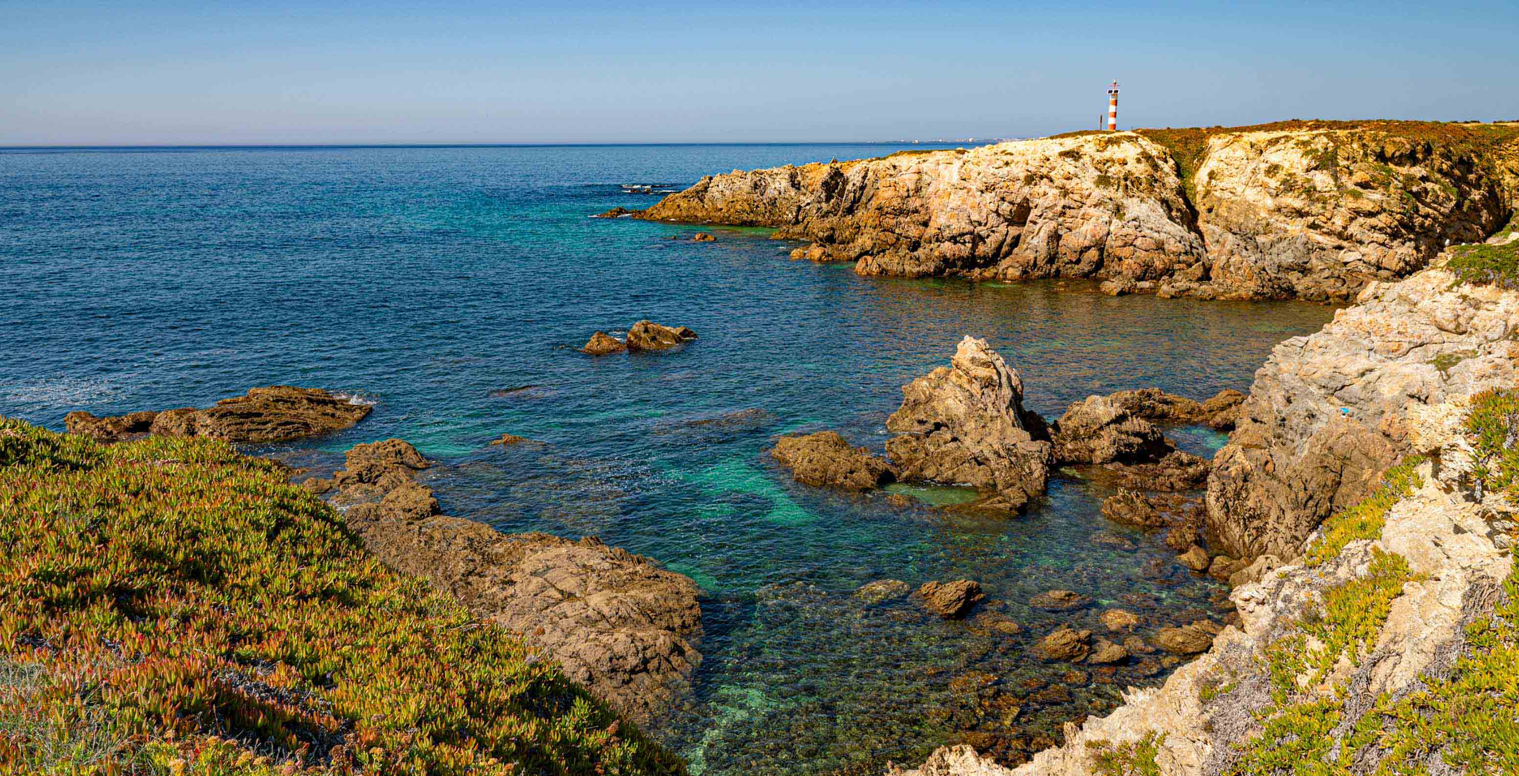 Falésias rochosas e mar azul junto ao Pestana Porto Covo Village, com um farol ao fundo