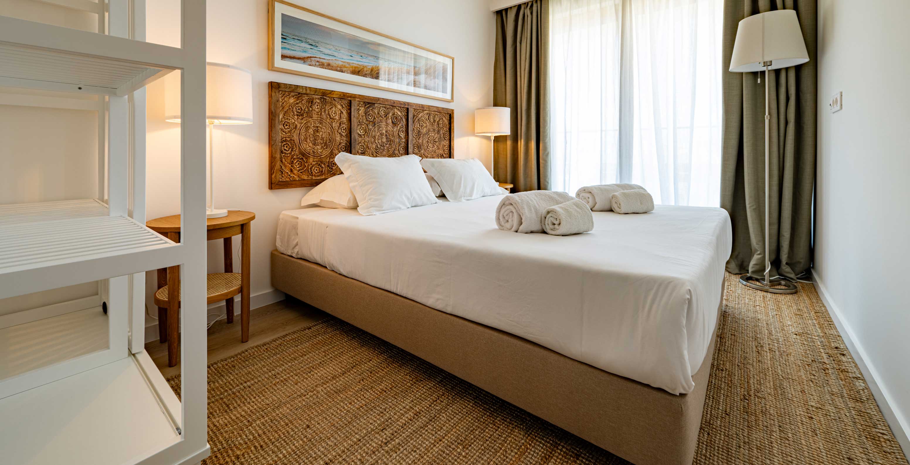 Quarto com cama de casal, cabeceira entalhada em madeira, candeeiros e luz natural no Pestana Porto Covo Village