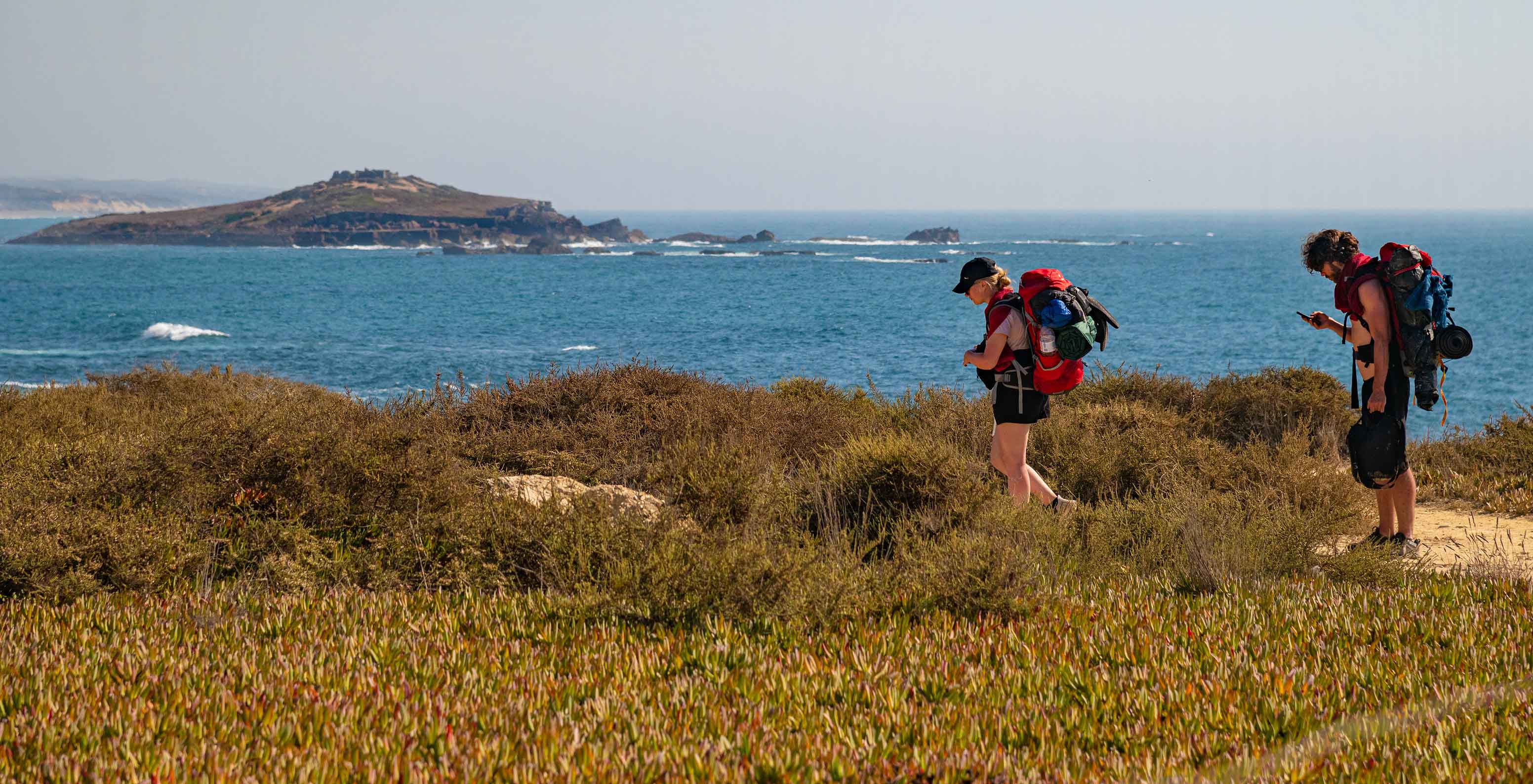 Trilheiro e trilheira com mochilas caminhando por trilha costeira com vista para o mar e ilha ao fundo