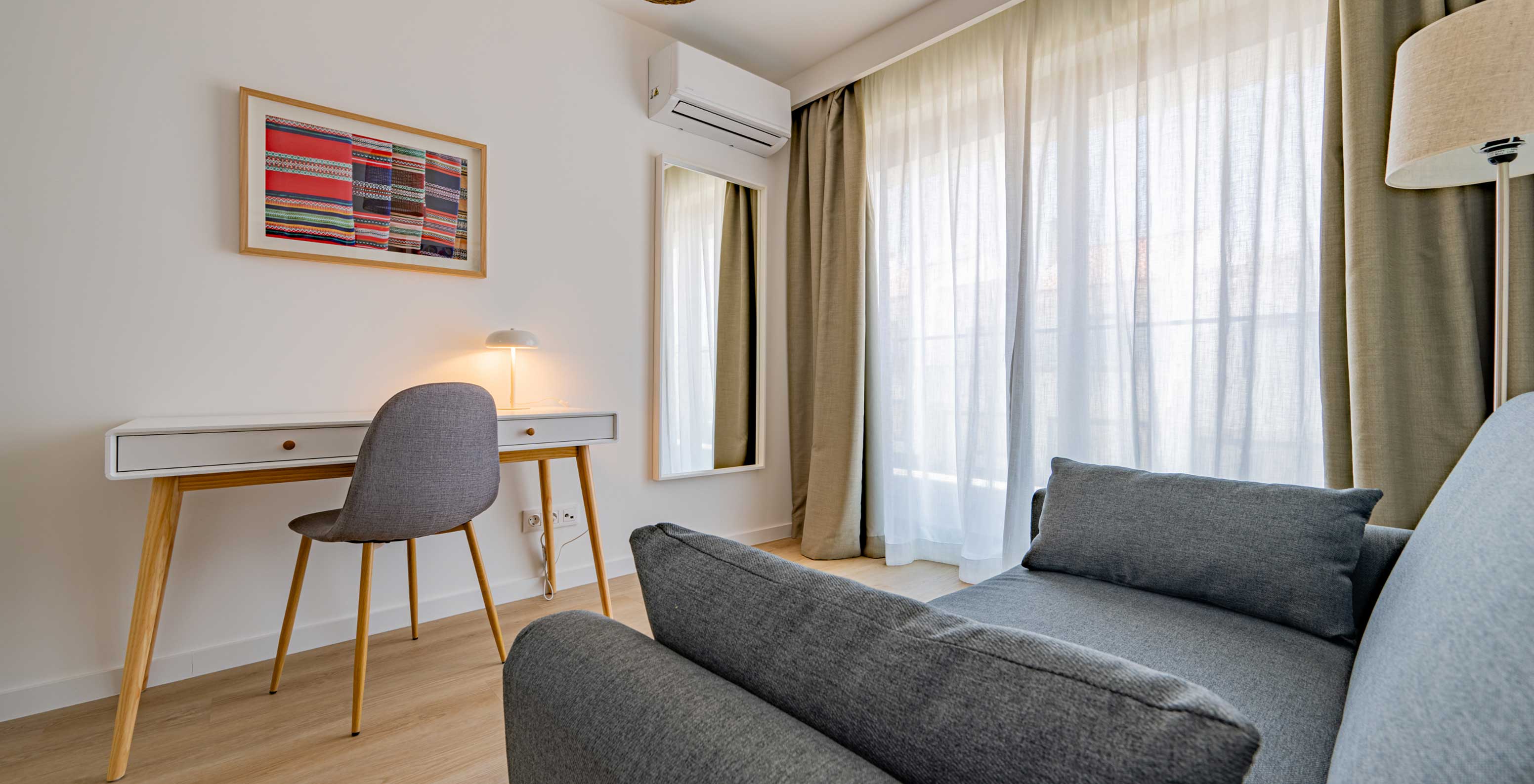 Área de trabalho no Pestana Porto Covo Village com secretária, cadeira cinzenta e sofá junto a cortinas claras