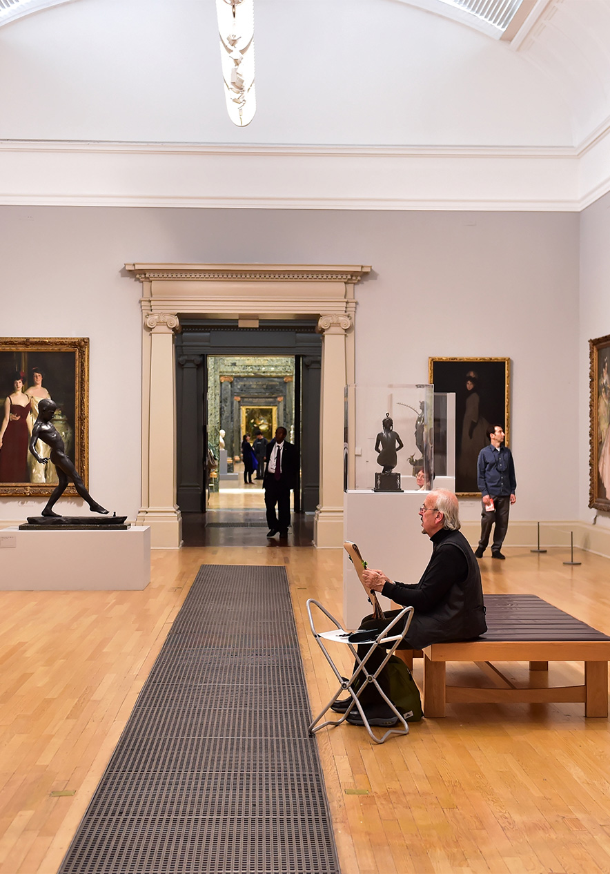 Room in a museum with various paintings and sculptures people walking wooden floor and white walls
