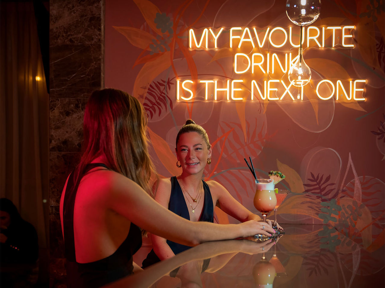 The Bridge Bar in London features two women at the counter with cocktails and neon lettering on the wall