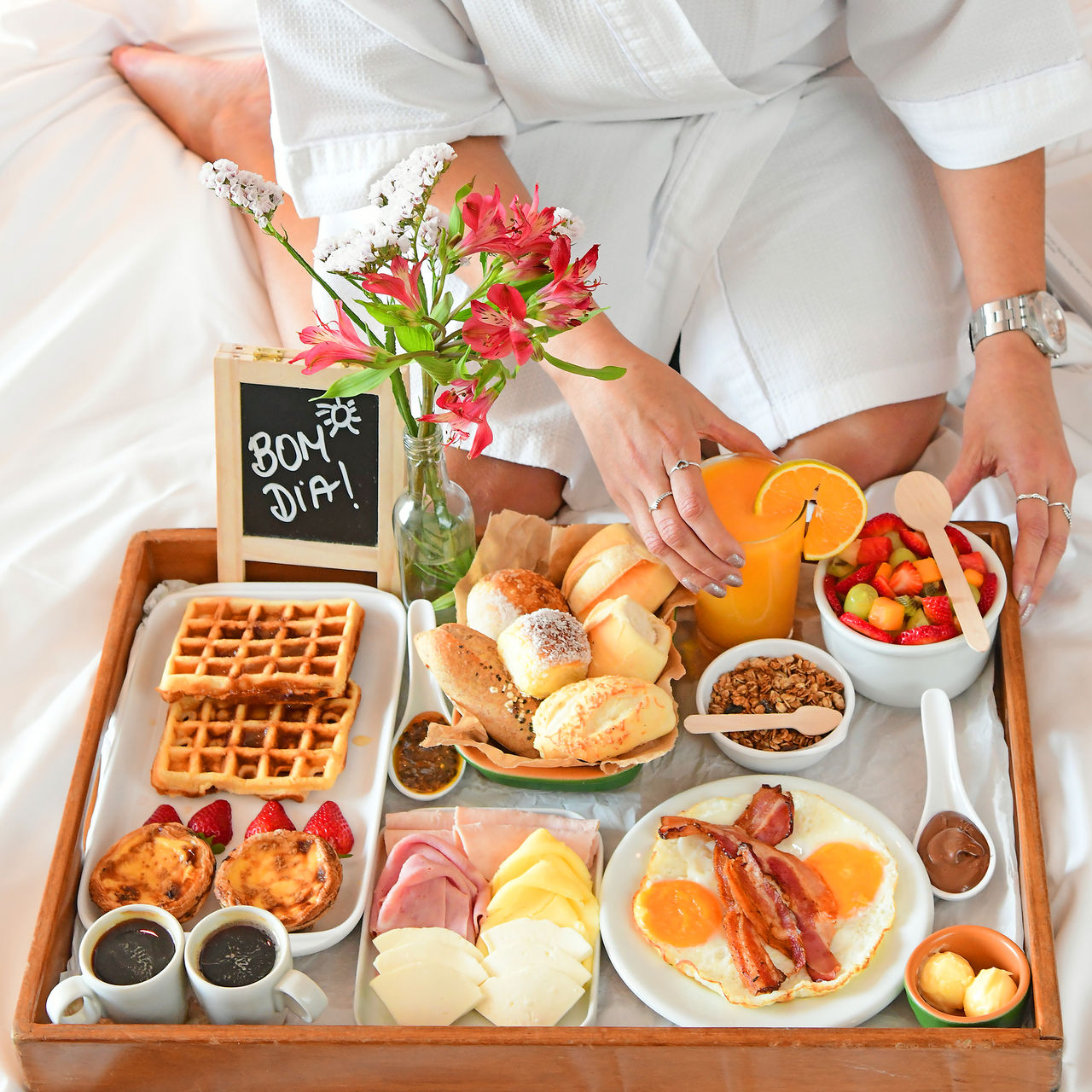 Girl in a robe having breakfast in bed, with a tray of bread, juice, coffee, waffles, cheese, ham, fruit, and eggs